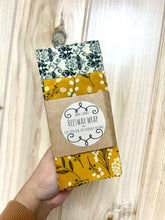 Load image into Gallery viewer, Hand holding a 2-pack of beeswax wraps. One is black and white with honeybees, the other is golden color. 
