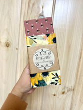 Load image into Gallery viewer, Hand holding a 2-pack of beeswax wraps. One wrap has floral print, the other has honeybees on a dark pink background.
